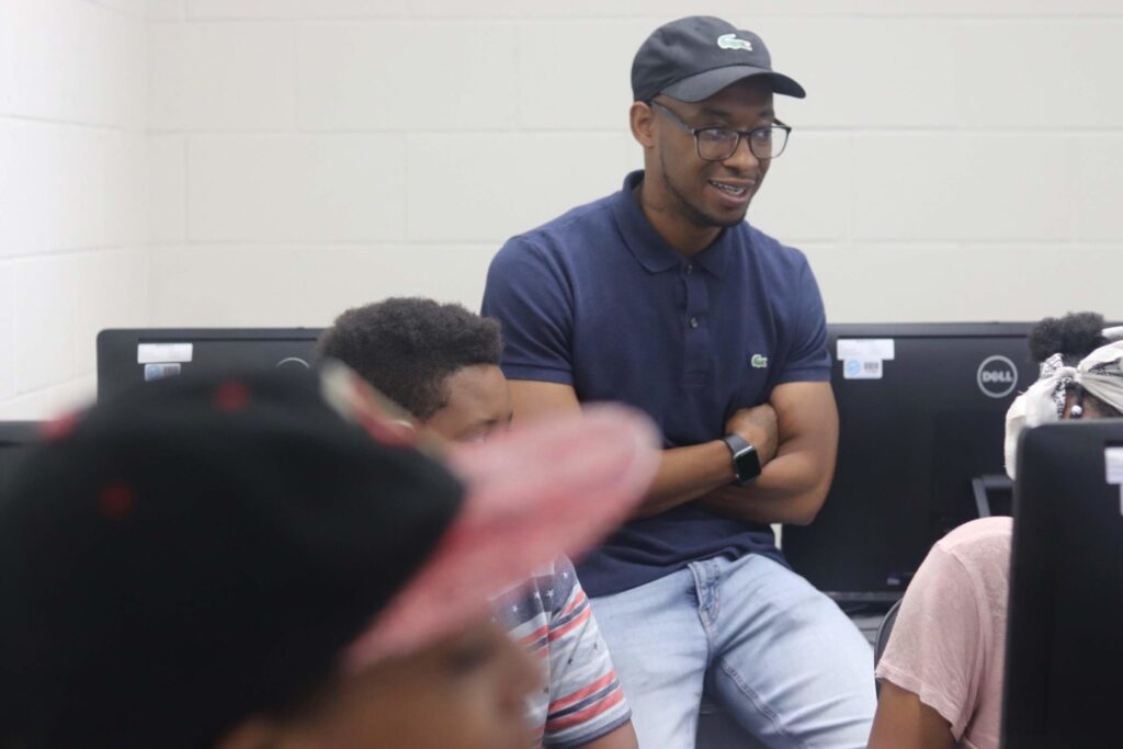 An adult smiles and talks to a group of kids seated at computers.