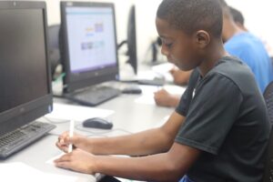 A young boy draws with markers while sitting at a computer screen.