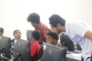 Two adults leaning down to talk to a row of children working on computers.