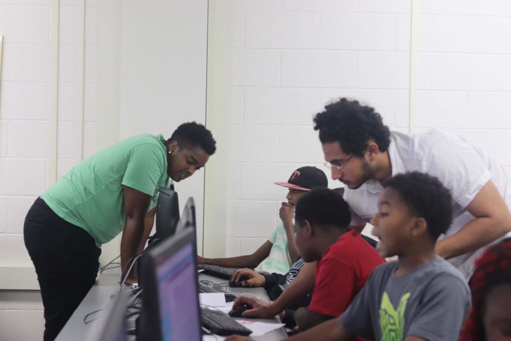 Two adults leaning down to talk to a row of children working on computers.