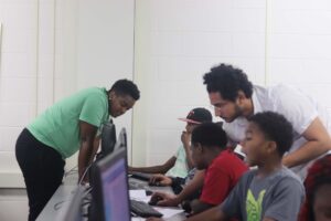 Two adults leaning down to talk to a row of children working on computers.
