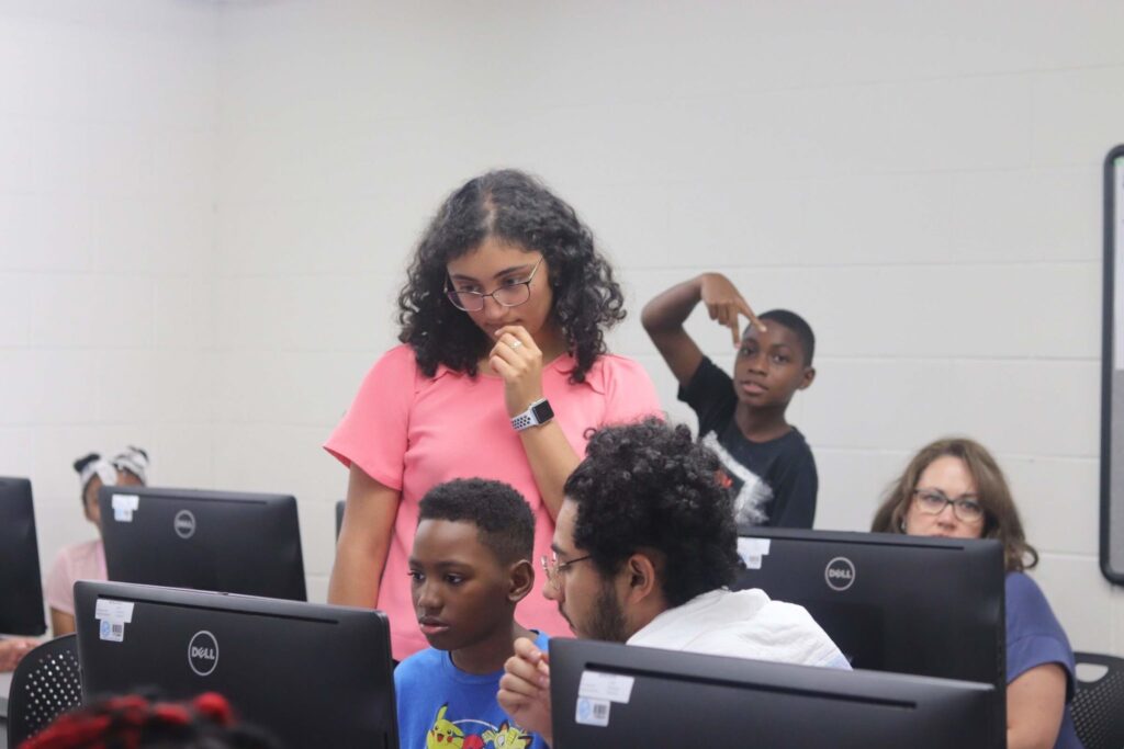 A child and two adults looking intently at a computer screen, with a child making a peace sign in the background.